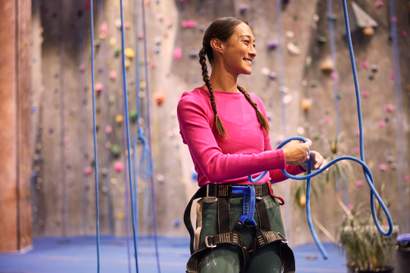 Mulher sorridente em academia de escalada indoor, usando equipamentos de segurança e corda, pronta para praticar a atividade física.
