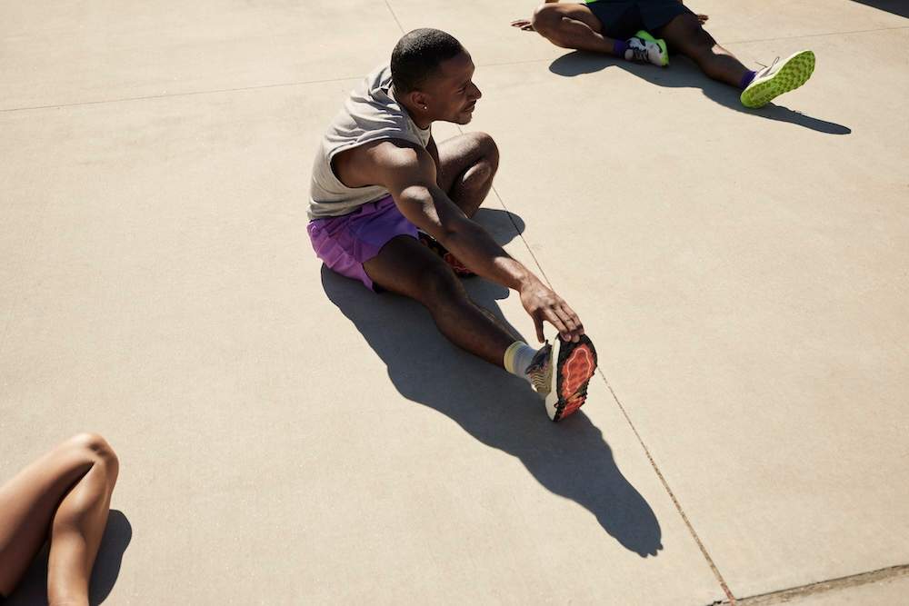 Homem em roupa esportiva se alongando ao sol sobre uma superfície de concreto, sentado e alcançando o pé; outras duas pessoas parcialmente visíveis também estão se alongando.
