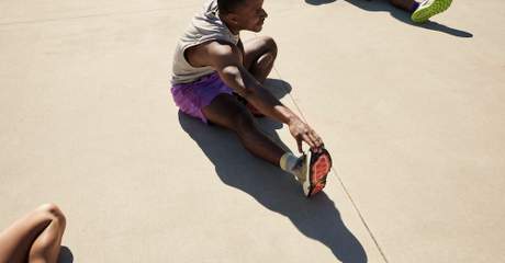 Homem em roupa esportiva se alongando ao sol sobre uma superfície de concreto, sentado e alcançando o pé; outras duas pessoas parcialmente visíveis também estão se alongando.