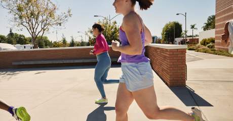 Três pessoas correm ao ar livre em um espaço urbano ensolarado, com árvores, prédios e um muro de tijolos ao fundo.