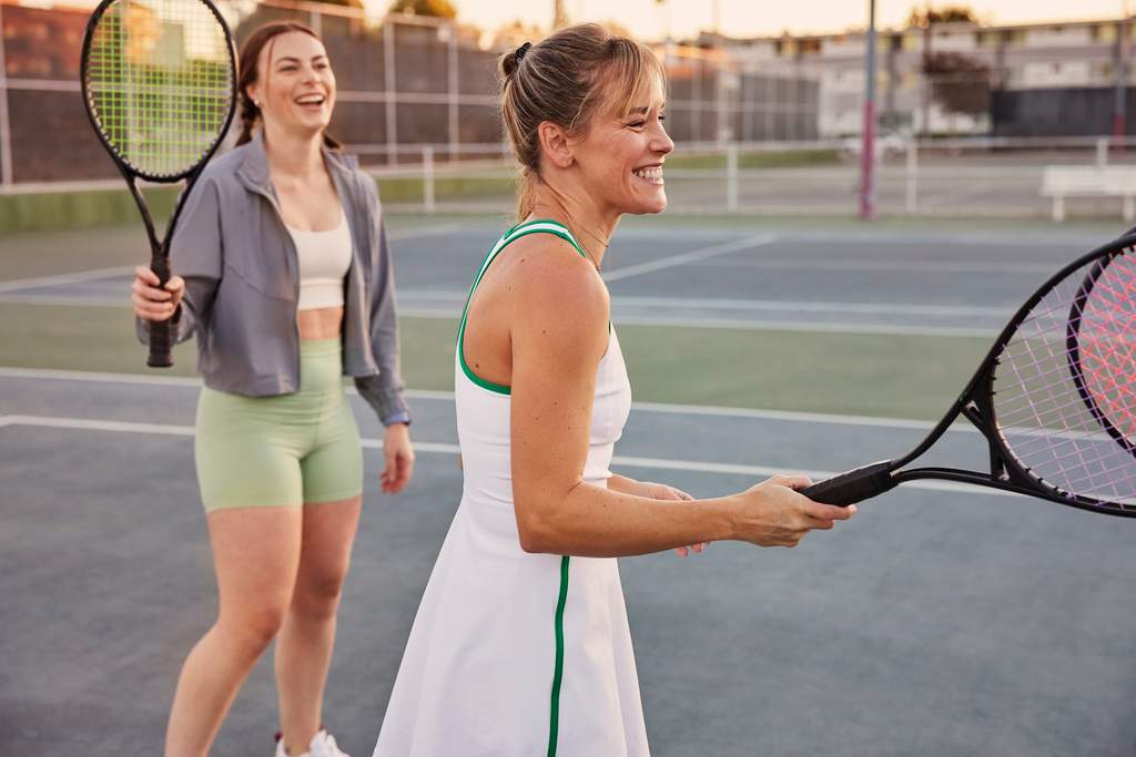Duas pessoas em uma quadra de tênis segurando raquetes, vestindo roupas esportivas e caminhando lado a lado após uma jogada.