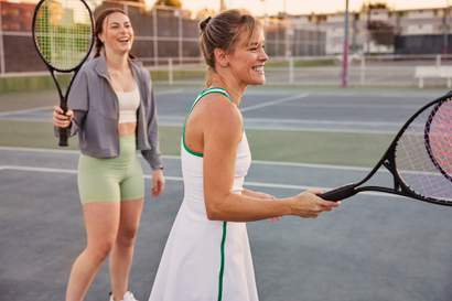 Duas pessoas em uma quadra de tênis segurando raquetes, vestindo roupas esportivas e caminhando lado a lado após uma jogada.