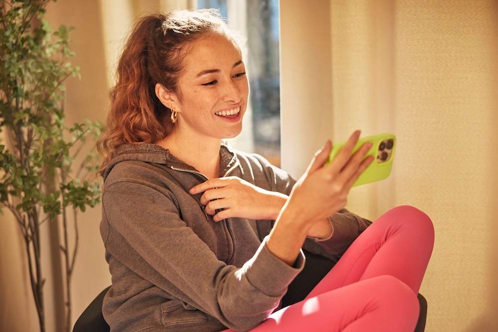 Mulher sorridente usando celular em casa, vestindo moletom marrom e calça rosa, em momento de bem-estar e relaxamento.