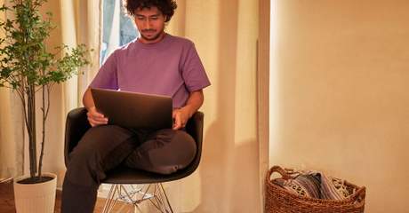Homem sentado em cadeira moderna, usando notebook no colo, em um ambiente aconchegante com planta decorativa e cesto de palha ao lado.