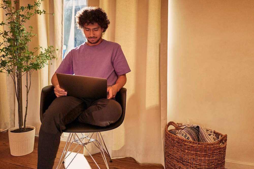 Homem sentado em cadeira moderna, usando notebook no colo, em um ambiente aconchegante com planta decorativa e cesto de palha ao lado.