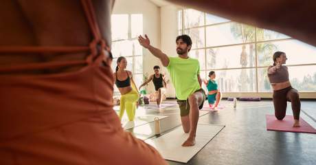 Grupo de pessoas praticando yoga em estúdio iluminado, realizando postura de alongamento e equilíbrio em tapetes de exercício.