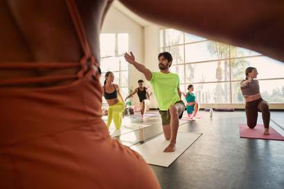 Grupo de pessoas praticando yoga em estúdio iluminado, realizando postura de alongamento e equilíbrio em tapetes de exercício.