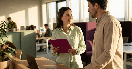 A woman holding a purple clipboard smiles while talking to a male colleague in a bright, modern open-plan office.