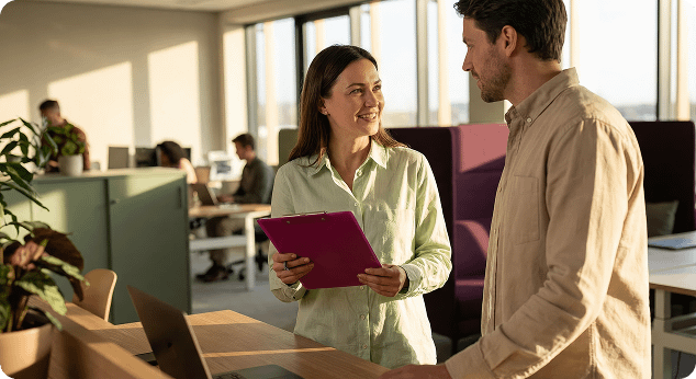 A woman holding a purple clipboard smiles while talking to a male colleague in a bright, modern open-plan office.