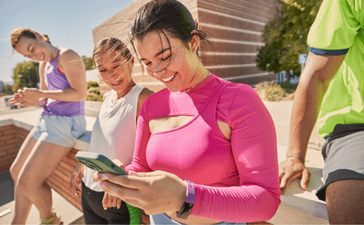 Quatro jovens em roupas esportivas relaxam ao ar livre, sorrindo e olhando para um celular nas mãos de uma mulher com blusa rosa vibrante.