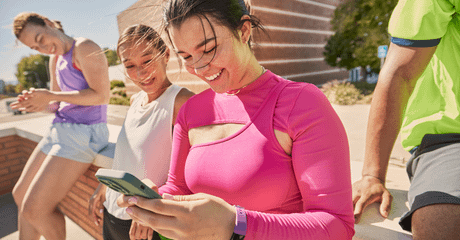 Quatro jovens em roupas esportivas relaxam ao ar livre, sorrindo e olhando para um celular nas mãos de uma mulher com blusa rosa vibrante.