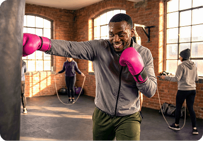 A smiling black man with pink boxing gloves punches a heavy bag in a sunlit brick gym.