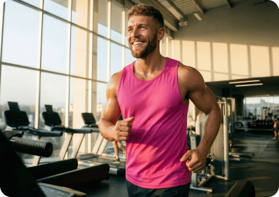A smiling white man in a pink tank top running on a treadmill in a sunlit gym