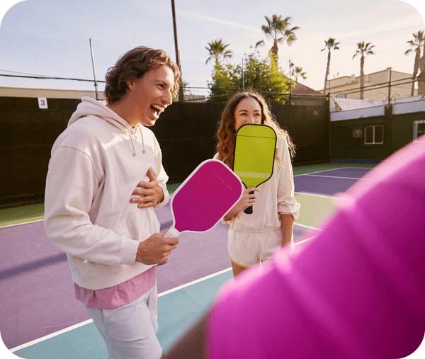 A man and woman laughing on a pickleball court while holding bright pink and lime green paddles.