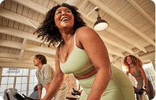 A smiling woman with curly hair leads a group in a bright, indoor cycling class under a beamed ceiling.