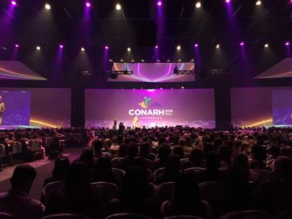 Auditório lotado com plateia assistindo a apresentação no palco do CONARH 2019, iluminado com luzes roxas e telão exibindo o logo do evento.