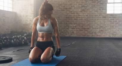 Mulher em treino de alta intensidade (HIIT) ajoelhada sobre colchonete em academia, usando roupas esportivas, após exercícios de força e resistência.