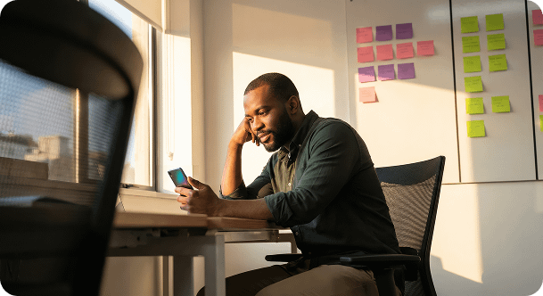 A man in a green shirt sits at a desk, looking at his smartphone in a sunlit office.