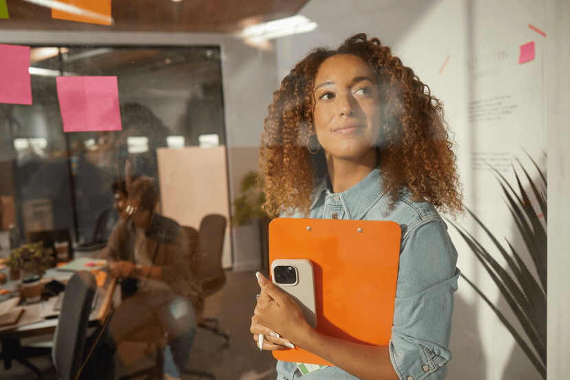 Pessoa em pé em frente a uma parede de vidro com post-its coloridos, segurando uma prancheta laranja e um celular, enquanto olha para o lado em um ambiente de escritório.