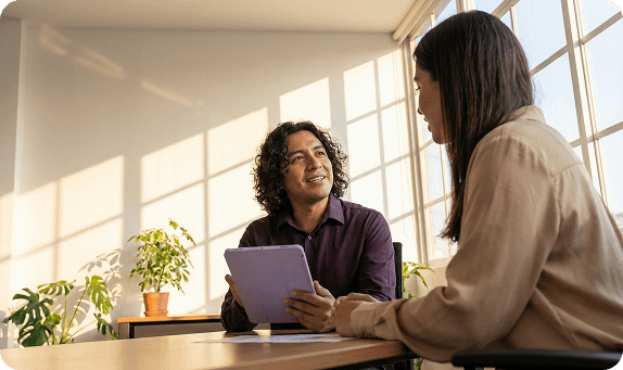 A man with curly hair holds a tablet while smiling and talking to a colleague in a sunlit office.