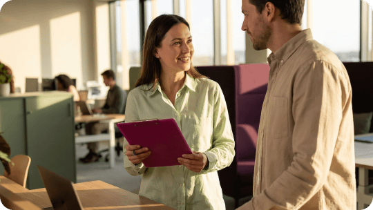 Mulher segurando prancheta rosa sorri enquanto conversa com colega em um escritório iluminado pelo sol.