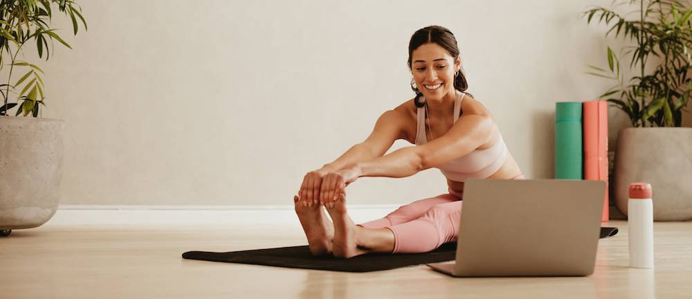 Woman sitting on the floor, wearing gym clothes, stretching, looking at a computer screen.