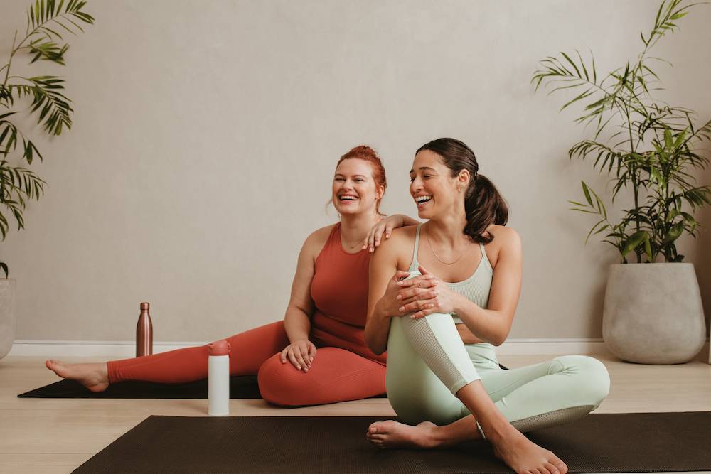 Two women sitting on the floor, wearing gym clothes, laughing.