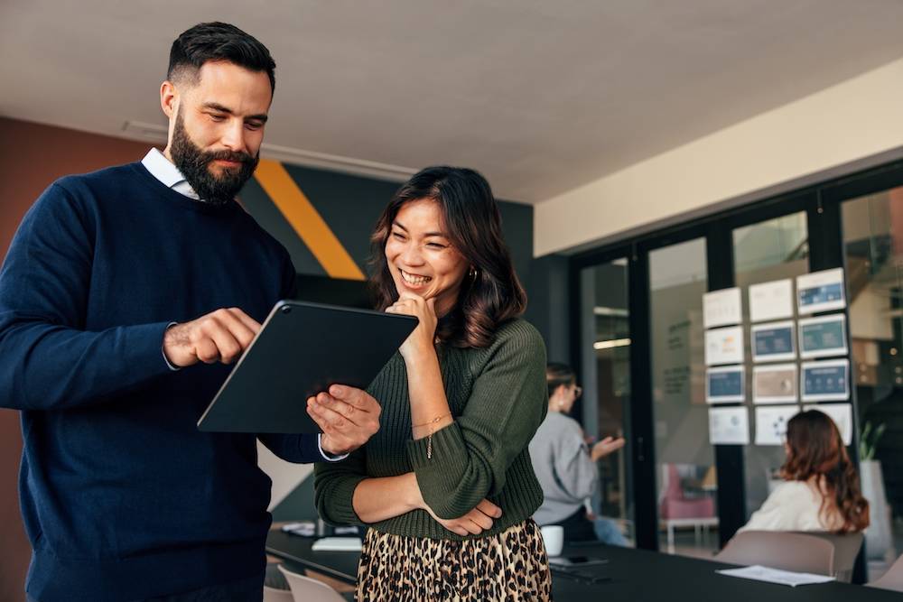 Two people, a man and a woman, at an office, looking at a tablet and smiling.