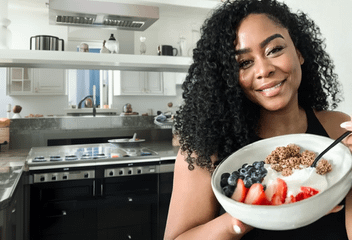 A smiling woman with curly hair holds a yogurt bowl topped with fresh strawberries, blueberries, and granola.