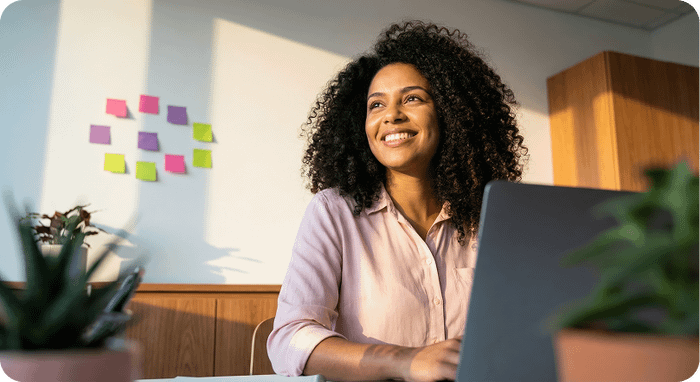 A smiling woman with curly hair sits at a laptop in a sunlit office with colorful sticky notes.