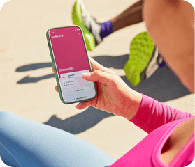 A person in workout gear holds a smartphone displaying the Wellhub app while sitting outdoors with other runners.