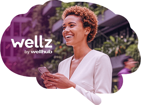 Logo Wellz by Wellhub ao lado de uma mulher sorridente segurando um smartphone em um ambiente ao ar livre.