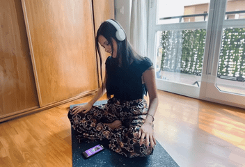 A woman sitting cross-legged on a yoga mat wearing headphones, using a smartphone app for guided mindfulness meditation.