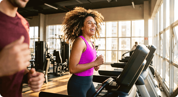 A laughing woman with curly hair runs on a treadmill in a bright gym during a sunset workout.