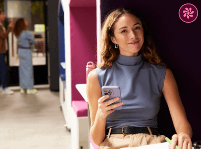 A smiling woman in a blue top holds a smartphone while sitting in a colorful, modern office meeting pod.