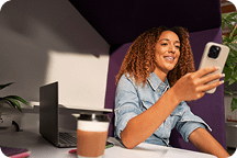 Mulher sorridente olha para o celular sentada em uma cabine de escritório com laptop e café à mesa.
