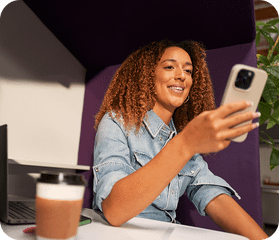 A smiling woman with curly hair uses a smartphone while sitting in a purple office privacy pod with a laptop.