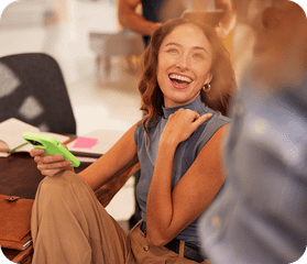 A smiling woman holding a smartphone, leaning against a colorful office booth with colleagues in the blurred background.